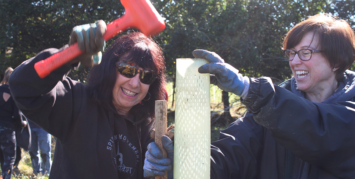 Two women smile as they pound tree stakes in a farm field.