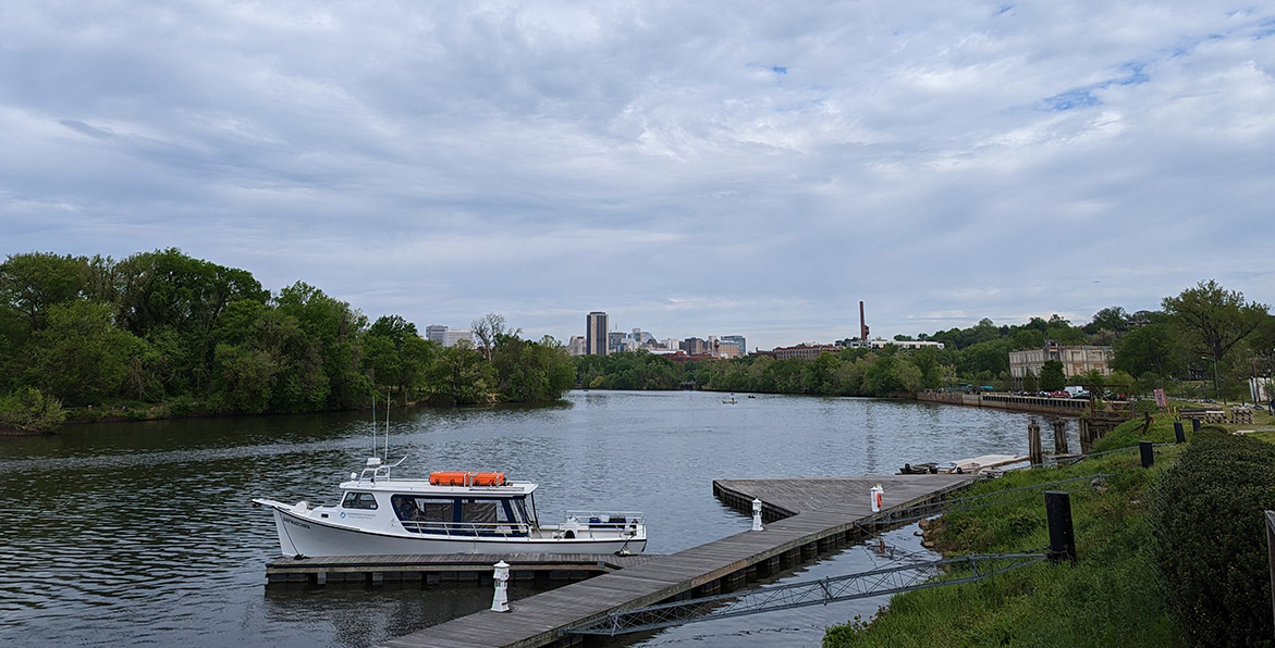 The CBF boat, Baywatcher, is docked on the James River, with the skyline of downtown Richmond in the distance.