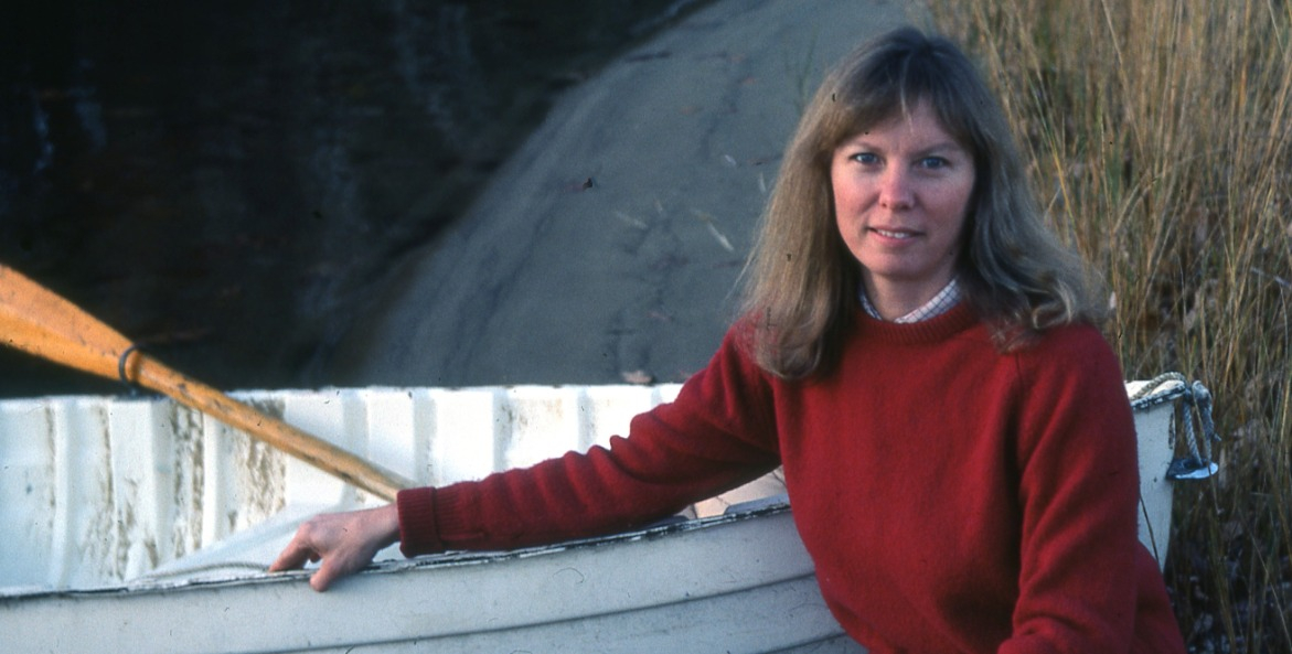 Woman in a red sweater kneeling along the water's edge next to a white row boat.