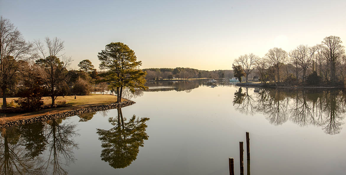 View from the bank of a calm creek with trees reflecting in the water.