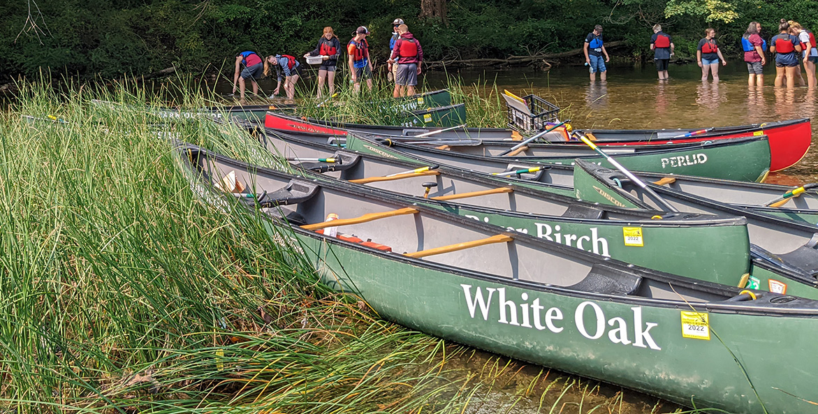 Students explore the shallows of a river with canoes featured in the foreground.