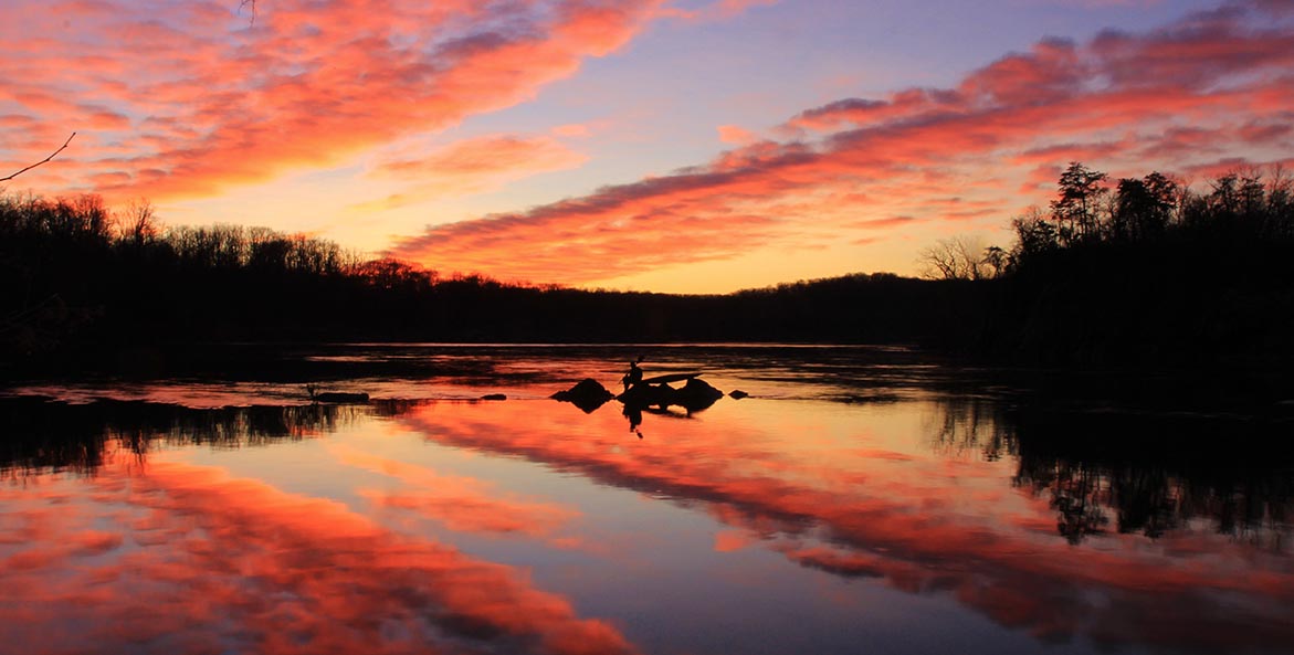 The rising sun illuminates the clouds orange over a still river and woodland.