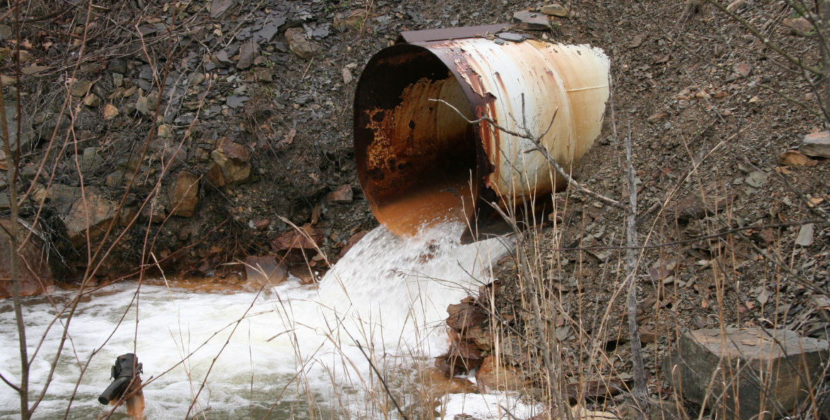 Acid mine drainage pours out of a large pipe and into a stream. The pipe and streambed are stained a rusty orange from the excessive iron deposits in the water.