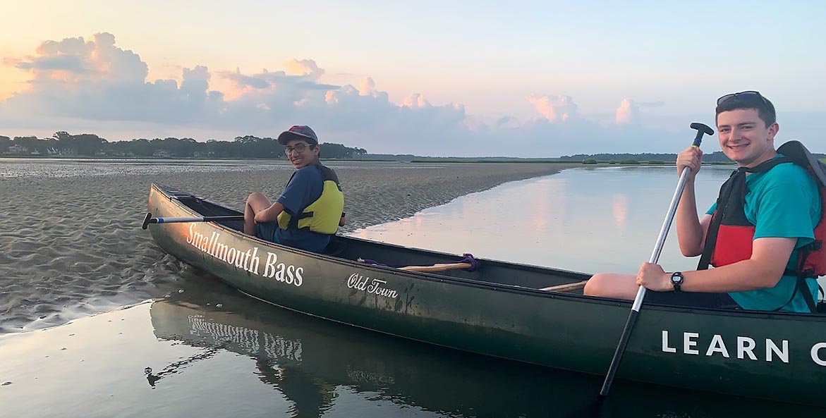 Two high school boys sit in a canoe with the name Smallmouth Bass pulled up on a sandy beach.