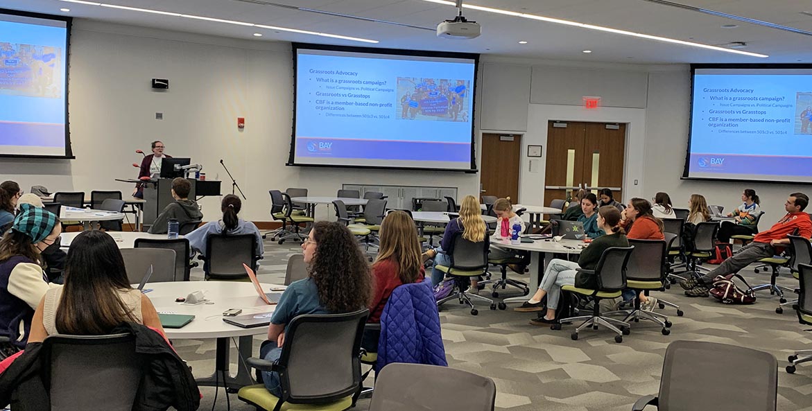 Students sit at round tables in a classroom with multiple presentation screens while an instructor makes a presentation.