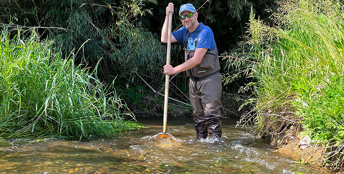 Man in high-waders stands in a stream with a net.