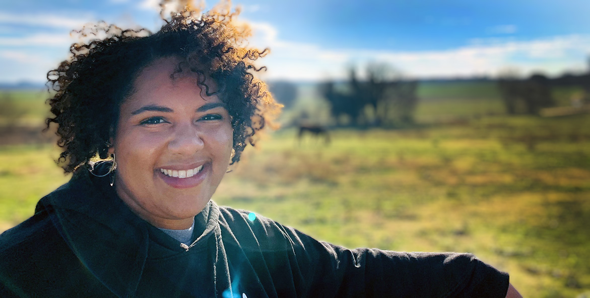 A Black woman smiles at the camera with a field and horse in the background.