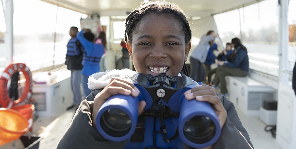 A young Black girl stands on the deck of a boat, holding a pair of binoculars and smiling at the camera.