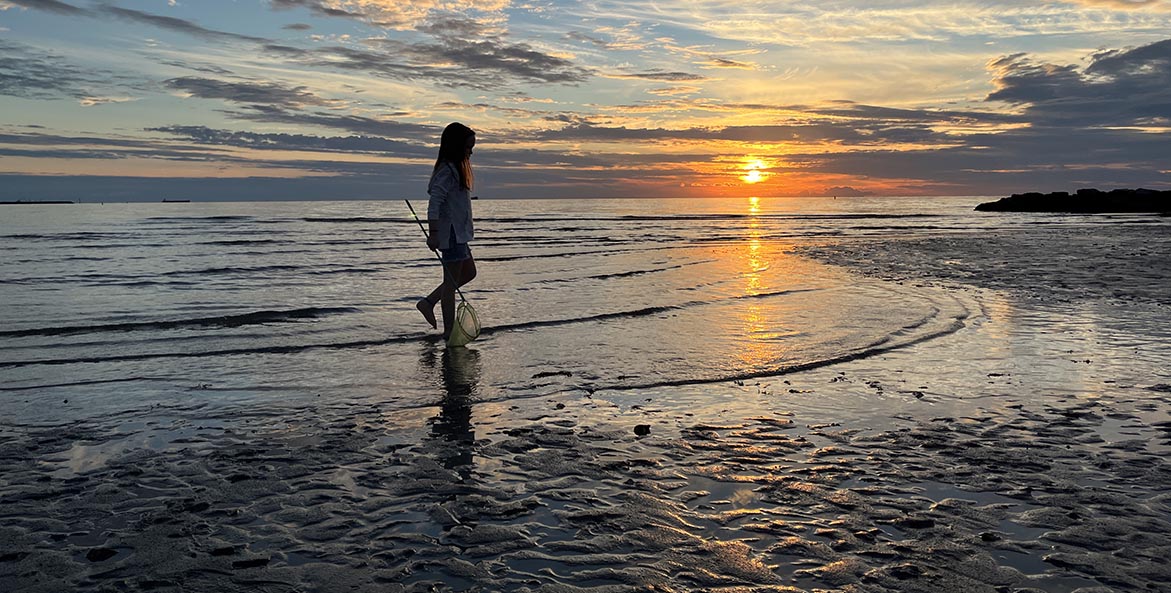 A young girl holds a fishing net and stands in the bay shallows during sunset.