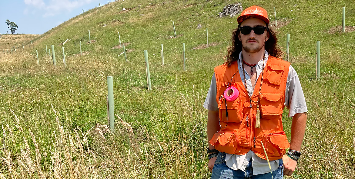 A young man in an orange vest and orange CBF cap stands in a filed of recently planted trees.