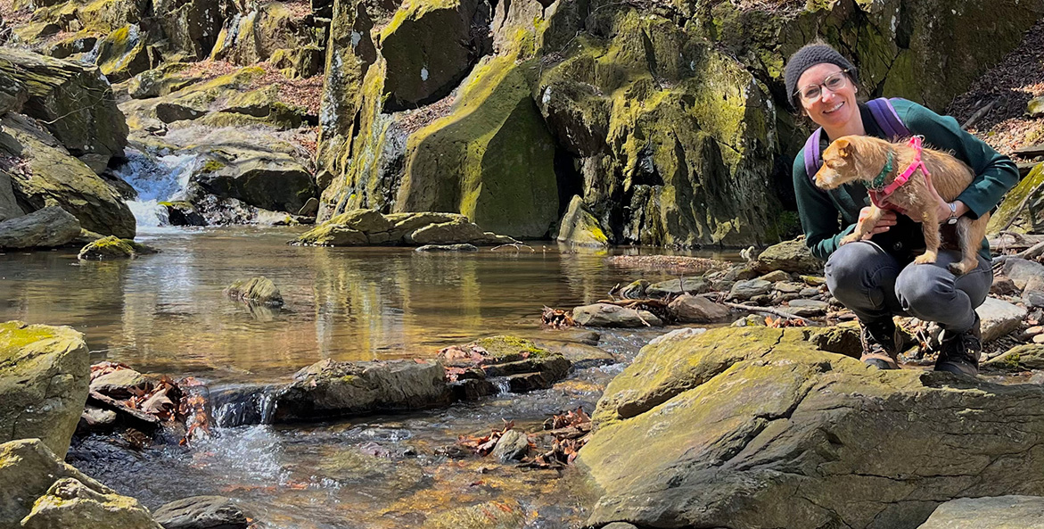 A woman crouches with her dog next to a rocky stream.