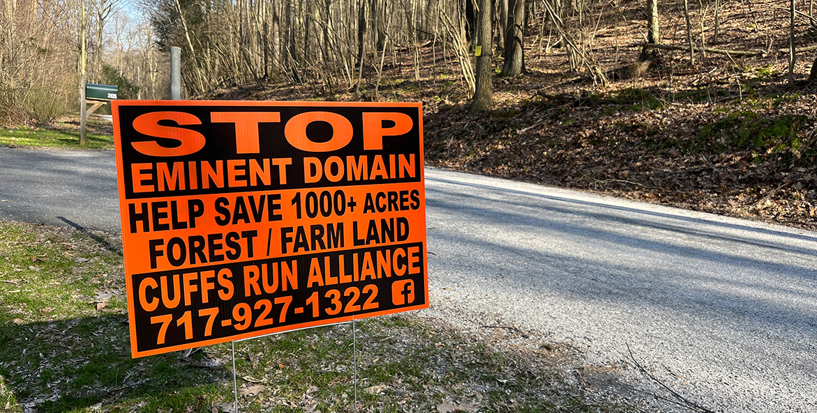 A roadside orange and black sign reads Stop Eminent Domain, Help Save 1000 plus Acres of Forest Farm Land.