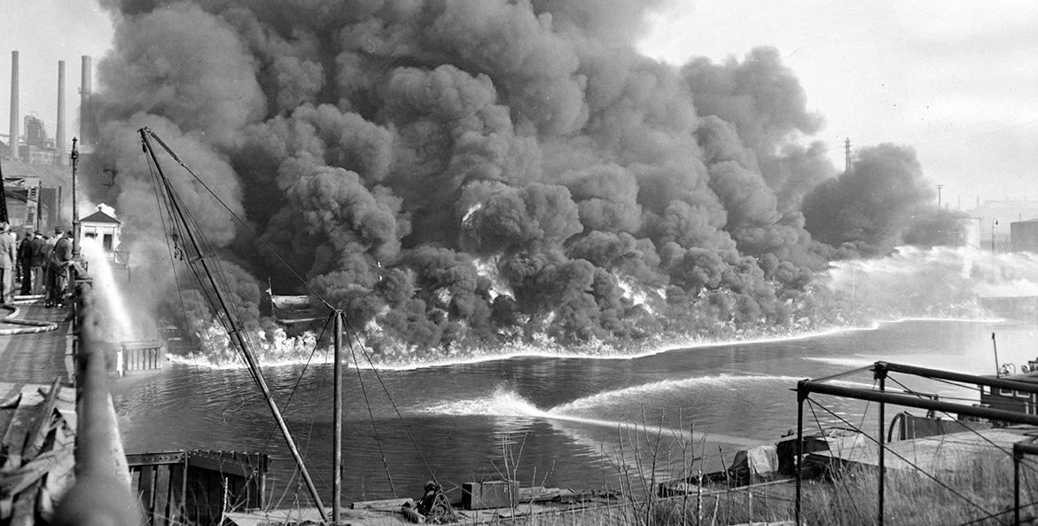 In this black and white photo, firefighters use water jets to corral a fire on the river to keep it from spreading.