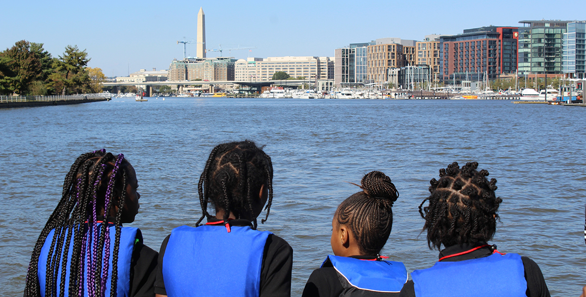The Washington Monument looms in the background as four students—with their backs to the camera—stand at the end of a boat looking at it across the water.
