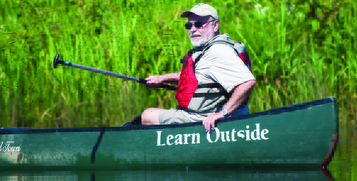 An older man with a white beard and mustach, sunglasses, and cap holds an oar while sitting in a green canoe with the lettering Learn Outside on its side.
