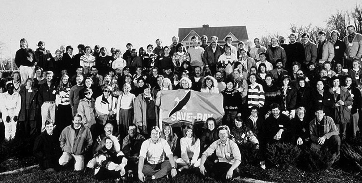 Older, black and white, photo of a large group of people holding a flag that reads Save the Bay.