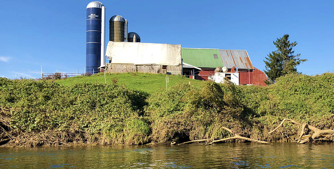 A farm with a red barn and blue silo sits on a hill adjacent to a creek.