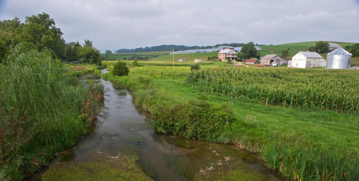 A stream runs through a lush green farm with barns, a silo, and rolling hills in the background.