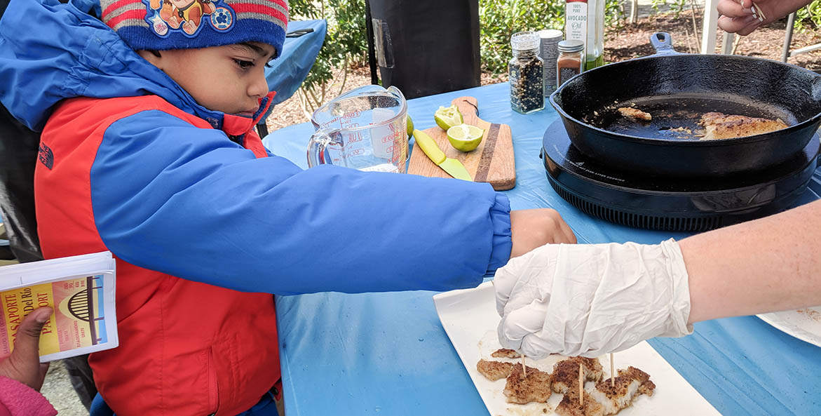 Young boy reaches for prepared fish on a demonstration table.