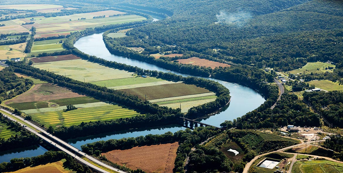 Thick buffers of trees line a river that winds through lush farm fields.