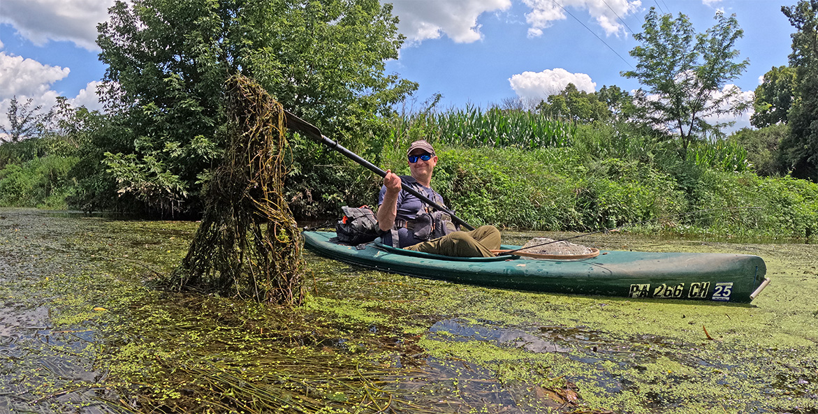 A man in a green kayak uses his paddle to lift a tangled mass of seaweed from the river's surface.