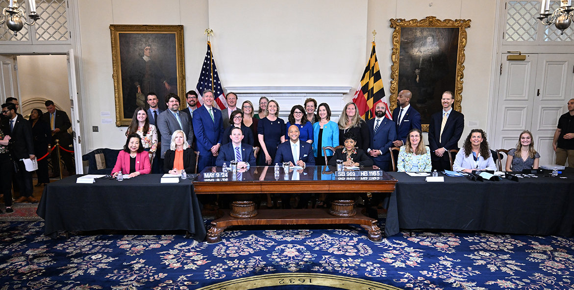 Many people including Gov. Moore smile behind a desk