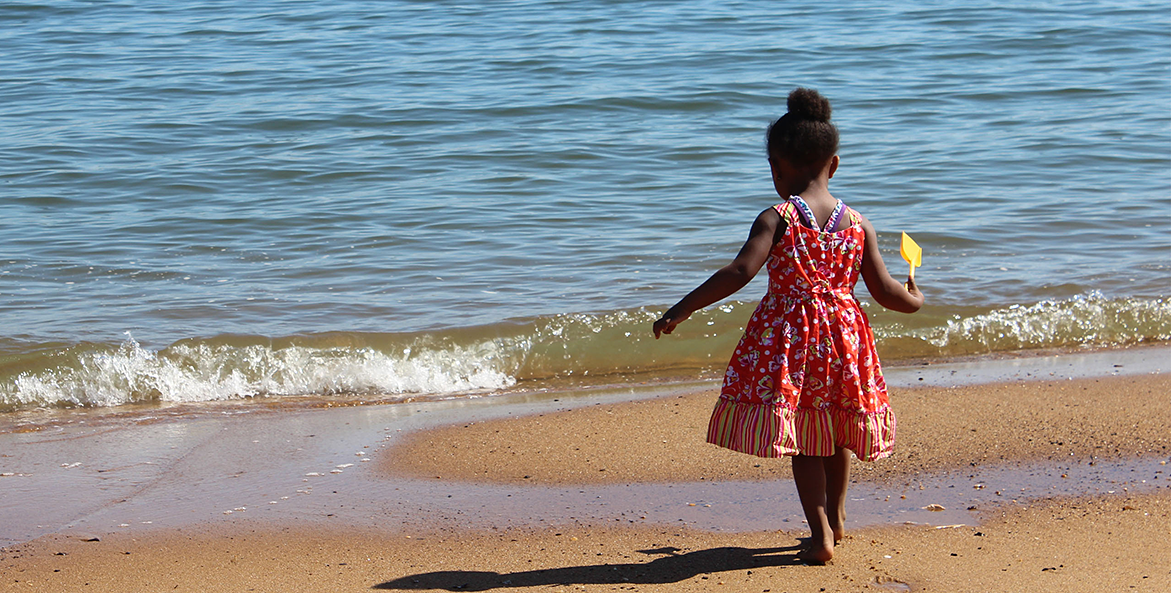 A little Black girl in a dress holds a sand shovel as she stands by the water's edge.