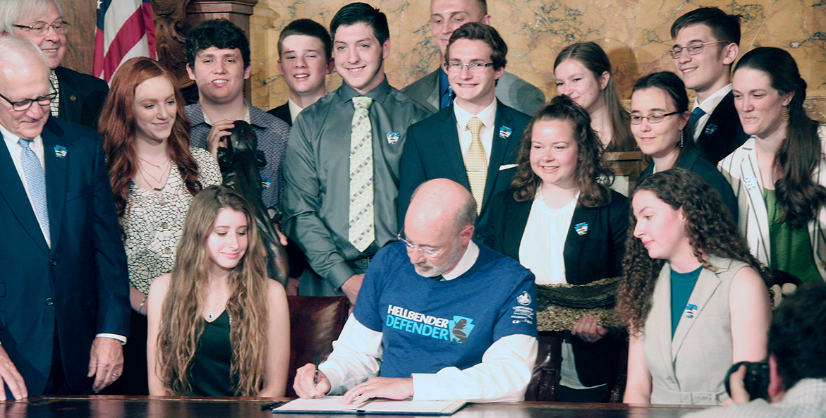 Gov. Wolf, center, wearing a blue Hellbender Defender t-shirt, is surrounded by students as he signs a bill.