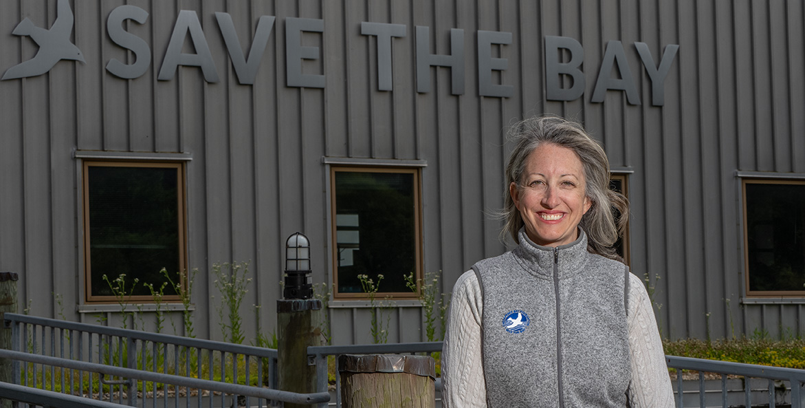 CBF President and CEO Hilary Harp Falk stands outside the front of the Philip Merrill Center on a fall day, the Save the Bay logo on the wall behind her.