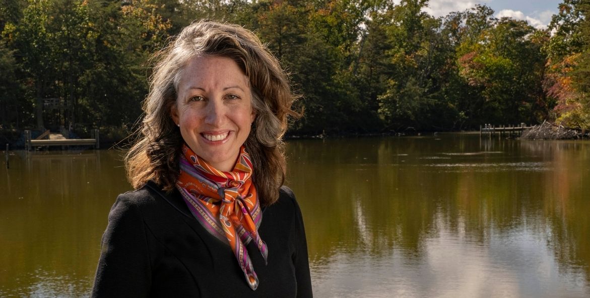 A smiling woman in a black shirt and red scarf stands in front of a wooded creek.