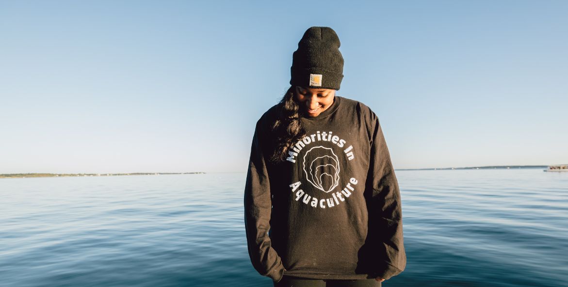 A woman wearing a knit cap and Minorities in Aquaculture sweatshirt smiles and looks down at the water.