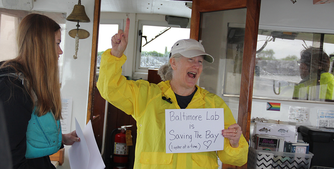 A woman in a yellow slicker smiles as she raises a finger in the air and holds a sign that reads Baltimore Lab is Saving the Bay 1 oyster at a time.