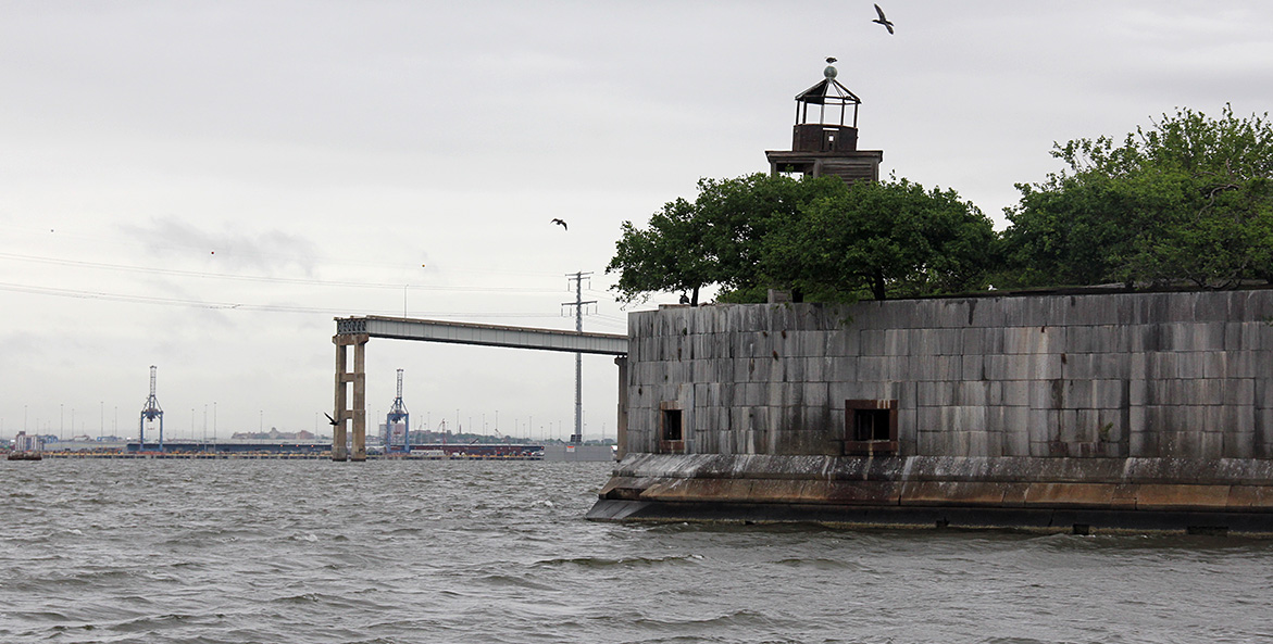 A gull flies over trees and a tower on a stone fort surrounded by water; the remains of a bridge in the background.