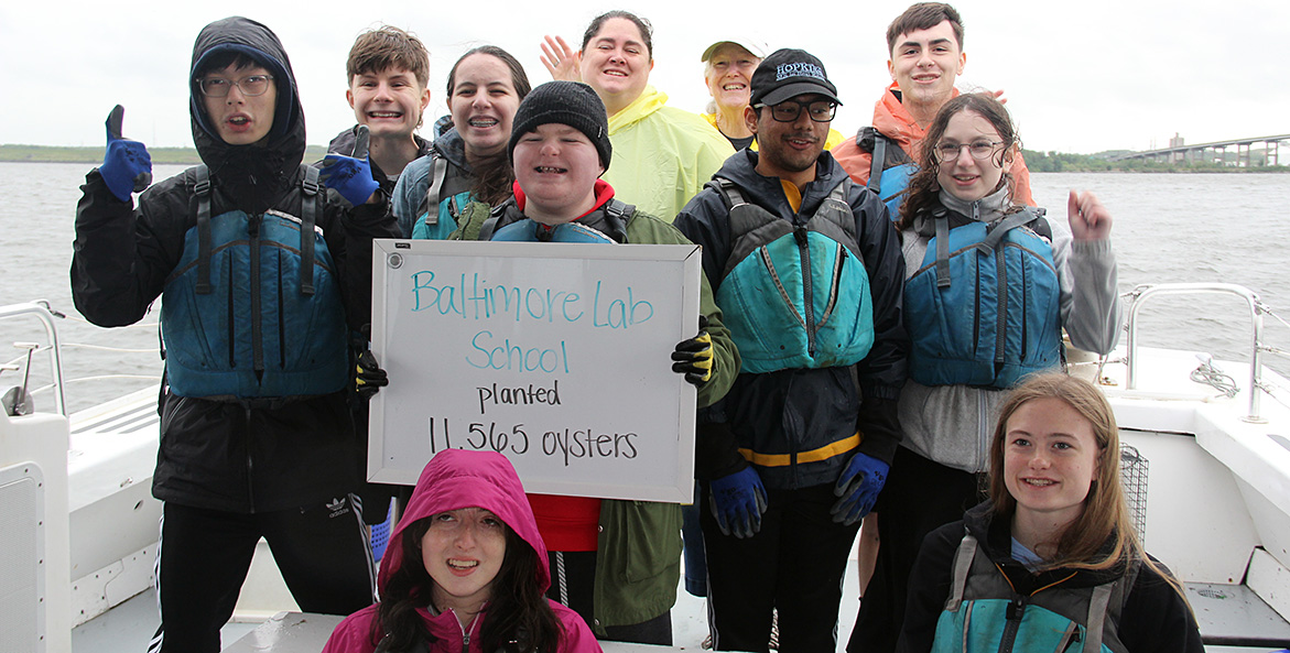 A group of smiling school students stand in a CBF boat, holding a sign that reads Baltimore Lab School planted 11,565 oysters.