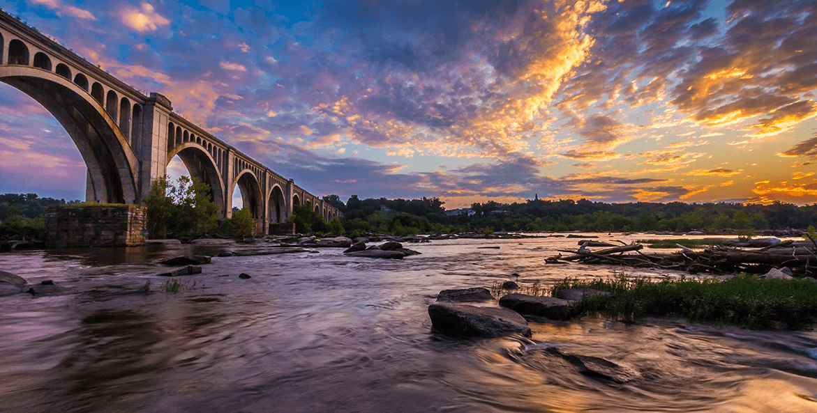 The summer sun rises and shines a rainbow of colors across the James River. The CSX A-line bridge crosses the river.