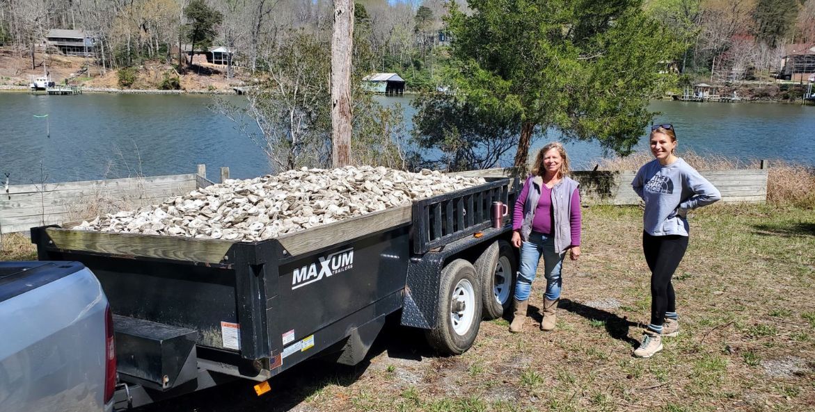 Two women stand next to a trailer filled with shells to be used for oyster restoration.
