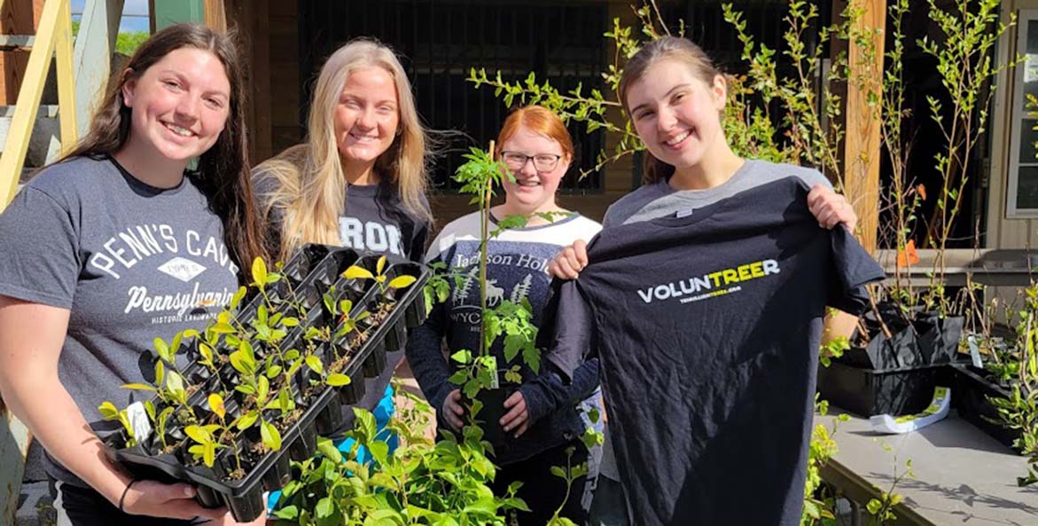 One adult and three students, two holding tree seedlings, one holding a tshirt standing with small trees.