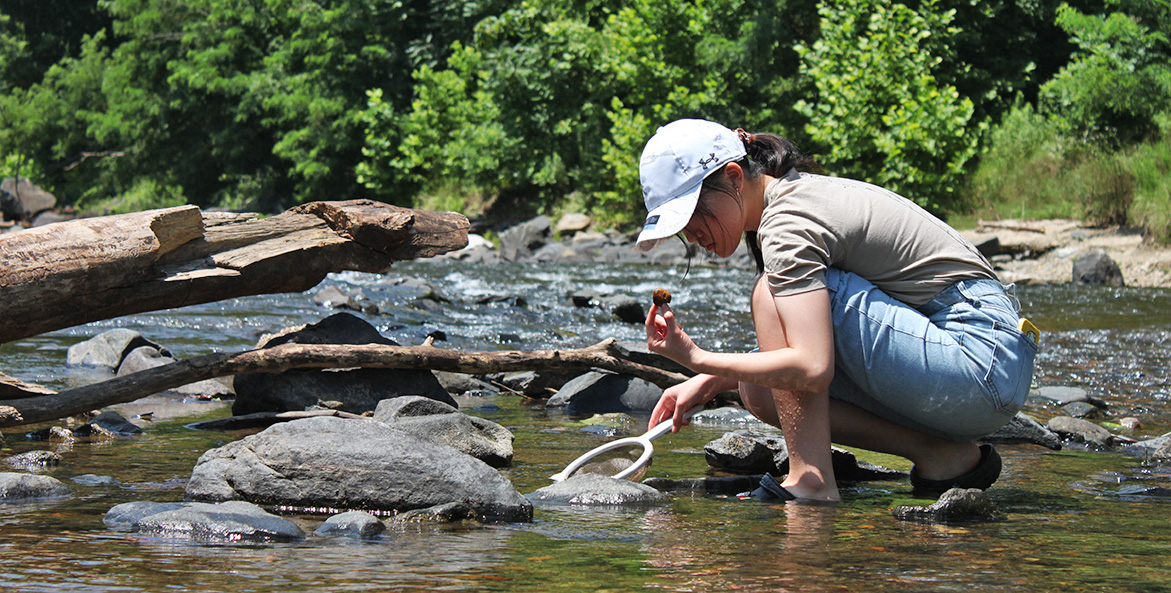 A teenager crouches in a stream, examining a round object from the stream floor.