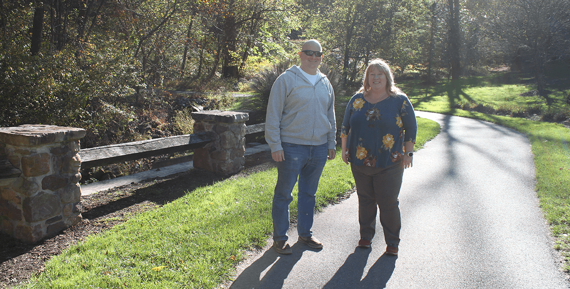 Man and woman stand on paved walking trail in front of grass and trees.