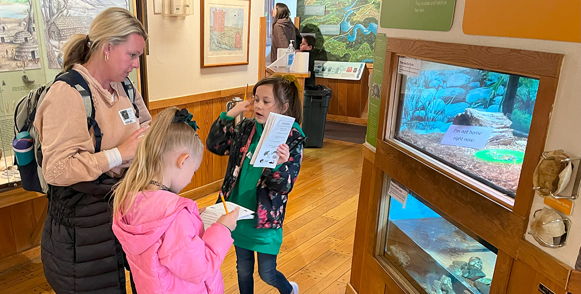 Two young students talk with an adult chaperone at a museum as they work on a study guide.