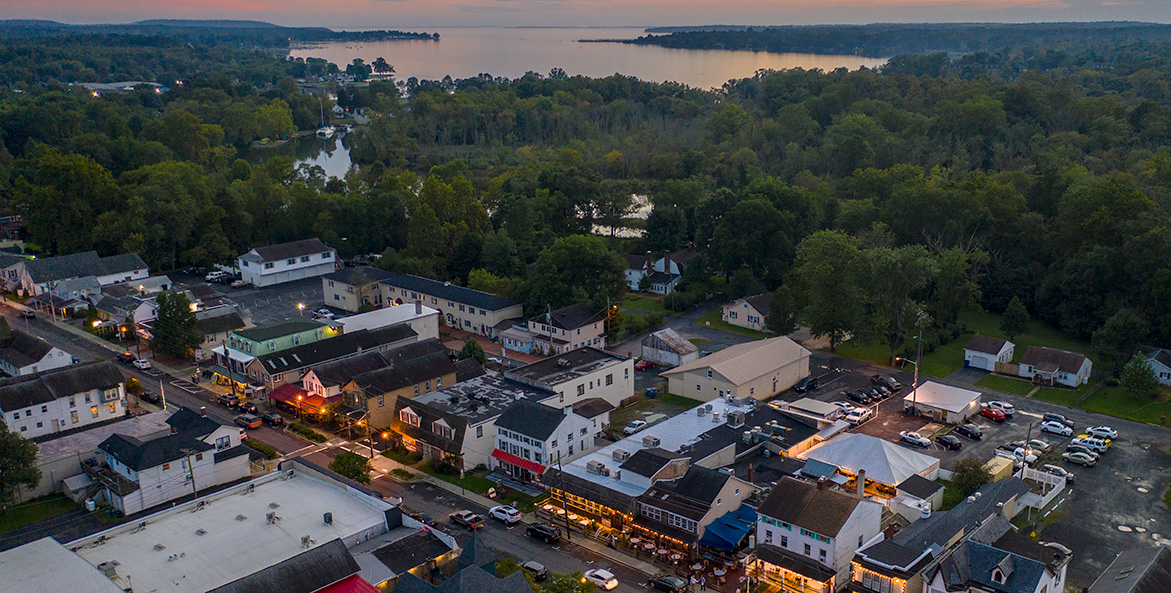 Streetlights and storefronts of small town mainstreet shine as dusk settles over the adjacent woodlands and river.