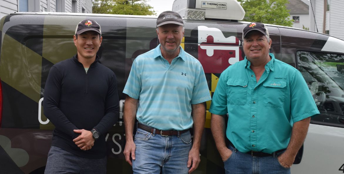 Three men stand in front of a van with a Maryland flag wrap.