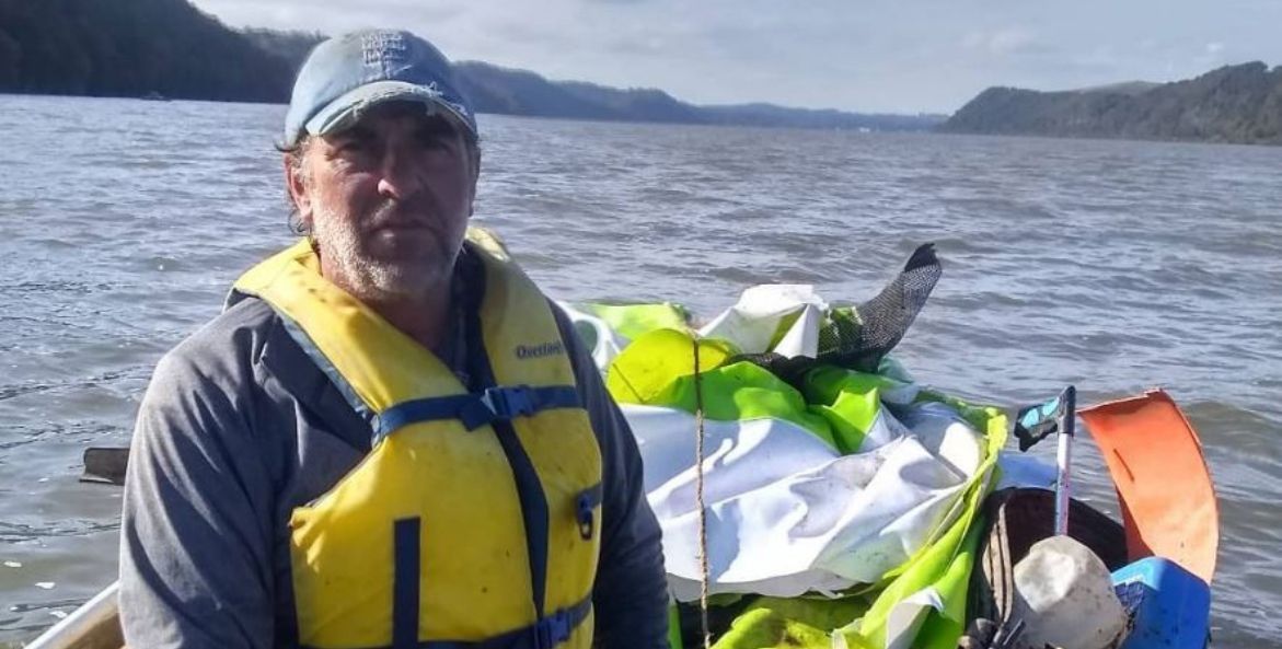 A man in a tattered, light blue baseball cap and yellow life vest sits in a boat with a pile of trash he's collected. Behind the boat, there is an expanse of trash-free water, surrounded by tree-covered mountains.