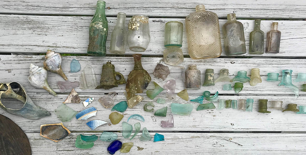 Sea glass, bottles, shells, pottery pieces lined up on wood slats.