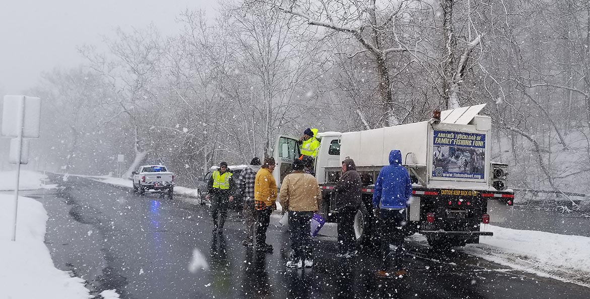 Snow falls as several men gather around a large white tanker truck parked on the side of a road next to a stream. 