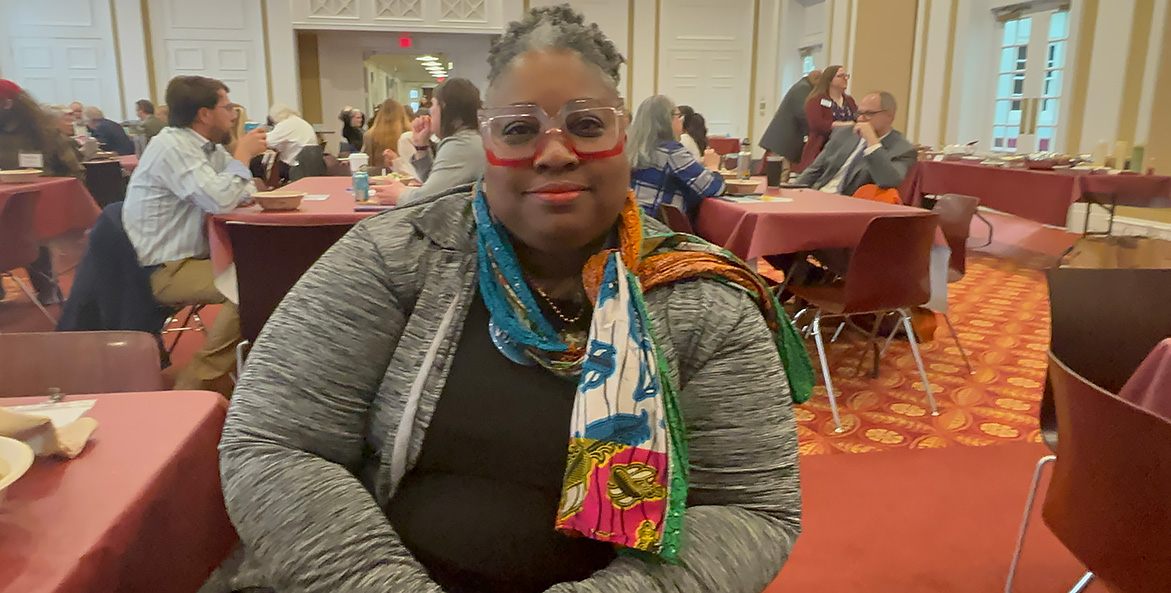 A Black woman with red-rimmed glasses and a colorful scarf sits in a conference room.