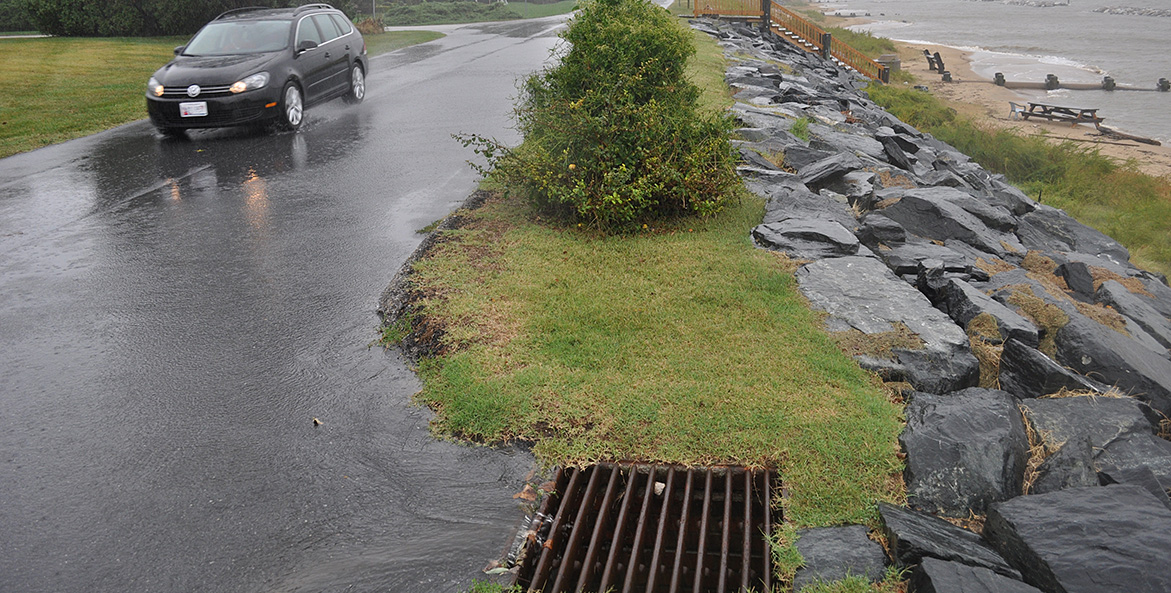 A car drives through the rain as stormwater flows into a storm drain.