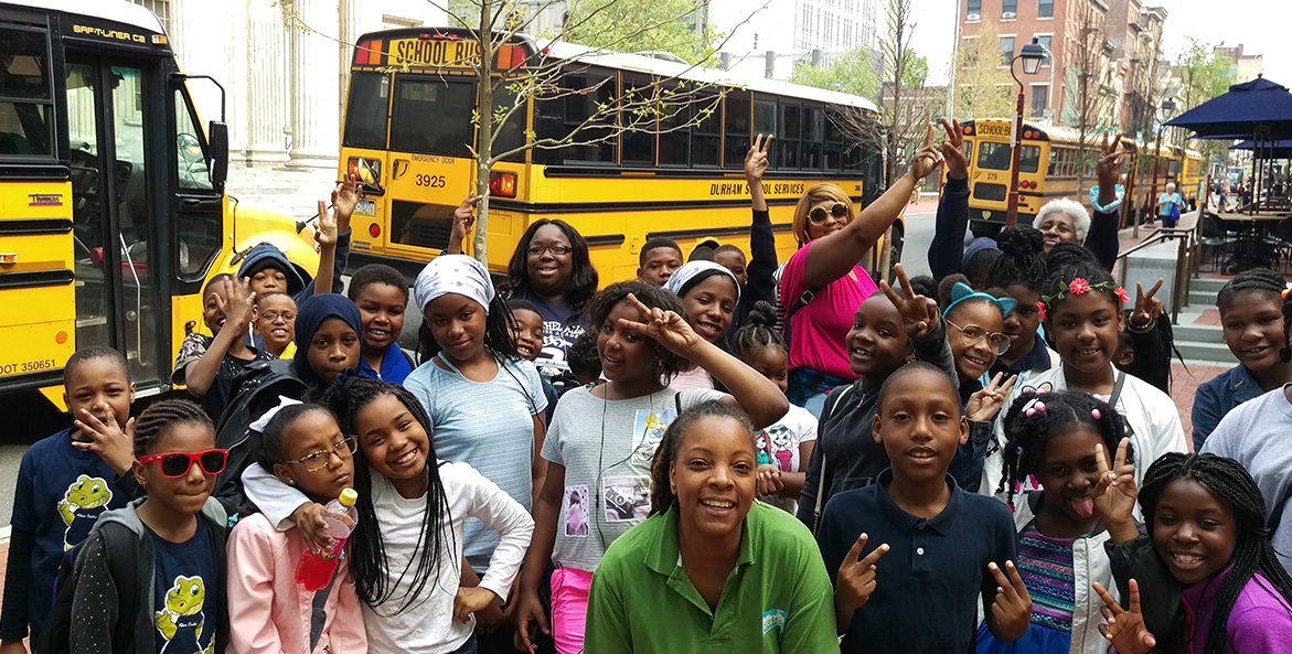 A crowd of students stand on a city sidewalk preparing to board school buses.