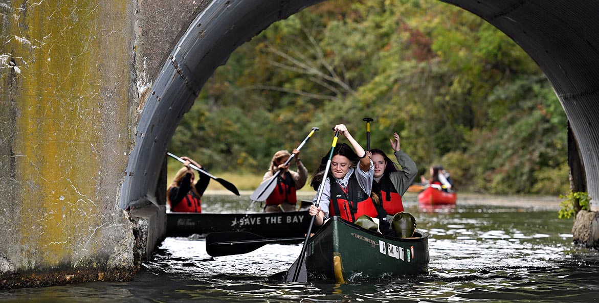 Students in red life jackets and green canoes paddle under a bridge.