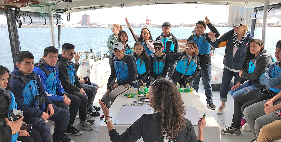 A large group of students are seated in the stern of a boat, raising hands and responding to an educator in the foreground.